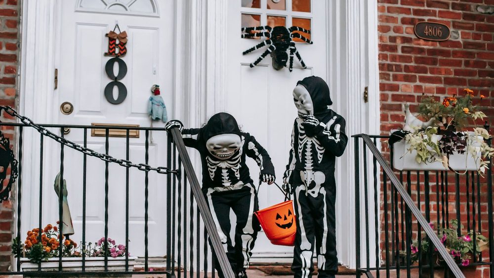 Twee kinderen in skeletkostuum met een pompoenemmer aan een versierde voordeur